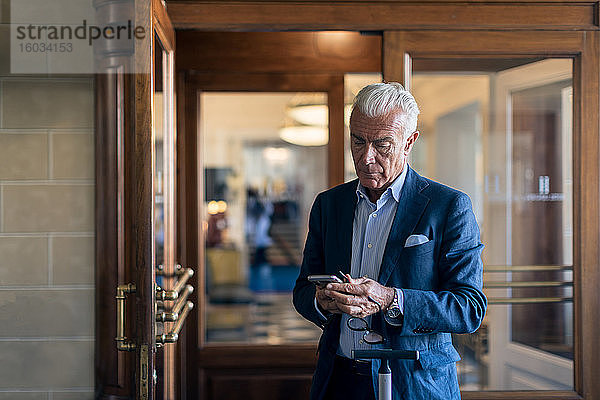 Ein älterer Geschäftsmann steht in der Hotellobby und schaut auf sein Handy.
