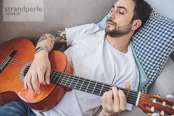 Auf dem Sofa liegender junger bärtiger Mann  der akustische Gitarre spielt.