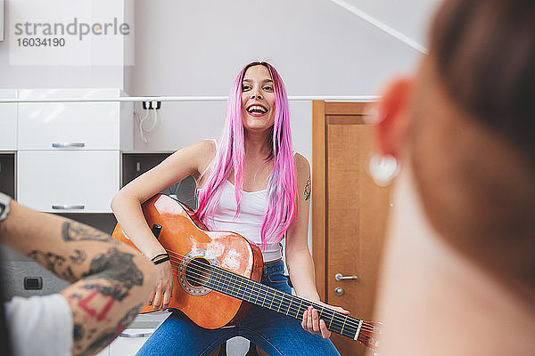 Junge Frau mit langen rosa Haaren  die in einer Küche sitzt und Gitarre spielt.