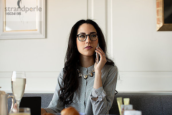 Frau mit langen schwarzen Haaren mit Brille  die an einem Restauranttisch sitzt und ein Mobiltelefon benutzt.