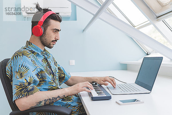 Junger bärtiger Mann  der an einem Tisch sitzt  Kopfhörer trägt  am Laptop tippt und auf der Tastatur spielt.