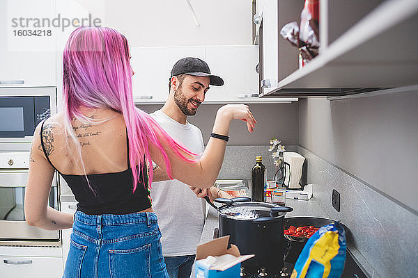 Junge Frau mit langen rosa Haaren und bärtigem Mann mit Baseballmütze steht in einer Küche und kocht.