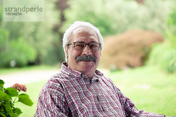 Porträt eines älteren Mannes mit Schnurrbart und Brille  der im Garten sitzt und in die Kamera lächelt.