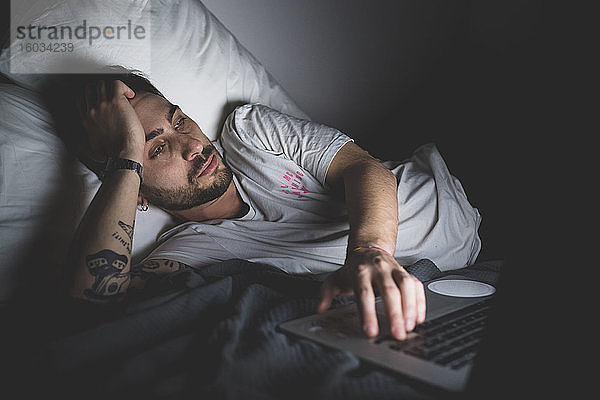 Bärtiger junger Mann  der nachts im Bett liegt und auf einen Laptop schaut.