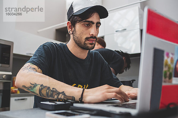 Junger bärtiger Mann mit Piercings  der an einem Tisch sitzt  eine Baseballmütze trägt und am Laptop tippt.