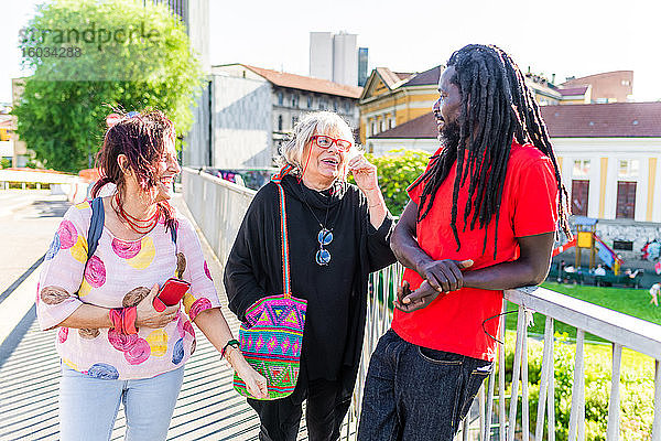 Schwarzer Mann mit Dreadlocks und zwei kaukasische Frauen stehen auf der Brücke  unterhalten sich und lachen.