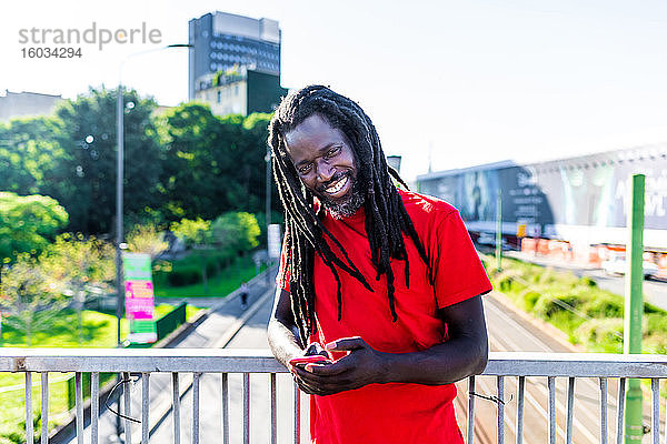 Porträt eines schwarzen Mannes mit Dreadlocks in rotem T-Shirt  der auf der Brücke steht und in die Kamera lächelt.