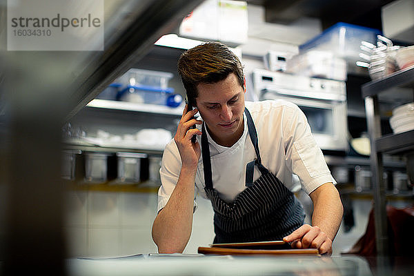 Junger männlicher Koch liest digitales Tablet und spricht über Smartphone in der Küche