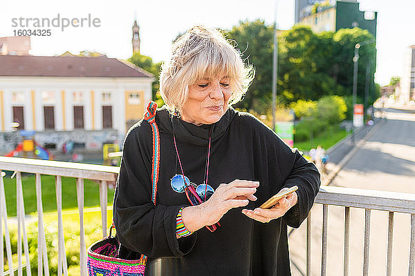 Ältere Frau mit blonden Haaren steht auf der Brücke und benutzt ein Mobiltelefon.