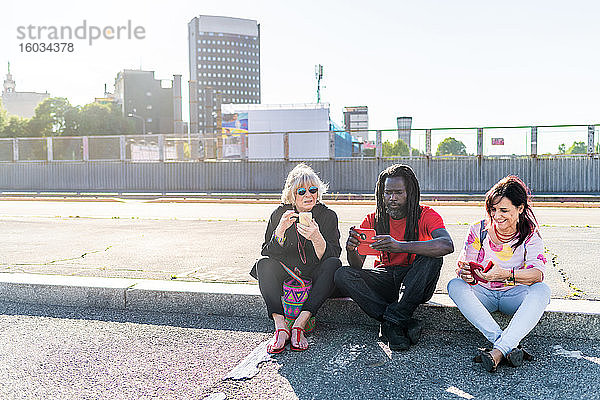 Schwarzer Mann mit Dreadlocks und zwei kaukasische Frauen sitzen auf dem Bürgersteig und überprüfen ihre Mobiltelefone.