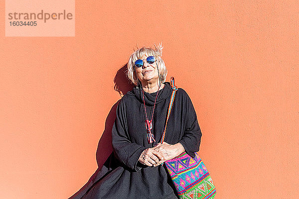 Porträt einer älteren Frau mit blonden Haaren und Sonnenbrille  die vor einer orangefarbenen Wand steht.