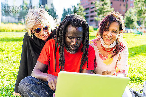 Schwarzer Mann mit Dreadlocks und zwei kaukasische Frauen sitzen auf dem Rasen und schauen auf einen Laptop.