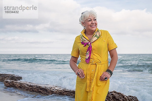 Porträt einer lächelnden Frau  die am Meer steht und ein gelbes Kleid trägt.