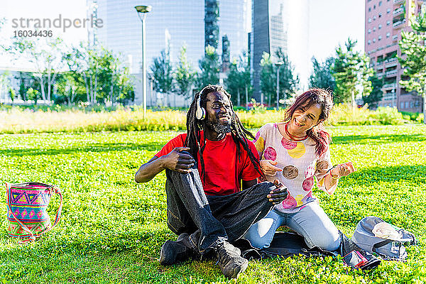 Schwarzer Mann mit Dreadlocks und kaukasische Frau sitzen auf dem Rasen und hören Musik.