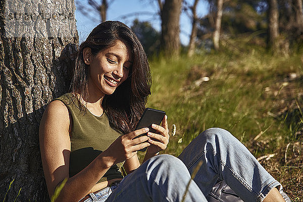 Porträt einer jungen Frau mit langen braunen Haaren  die unter einem Baum in einem Wald sitzt und ihr Handy überprüft.