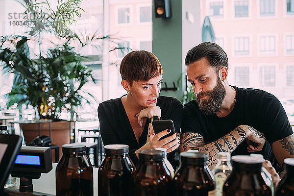 Bärtiger junger Mann mit braunen Haaren und Tätowierungen und junge Frau mit kurzen Haaren sitzt in einer Bar und schaut auf ein Mobiltelefon.