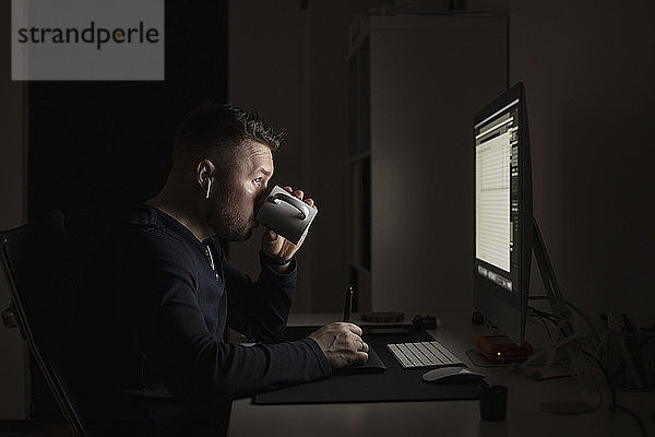 Mann mit Ohrstöpseln trinkt Kaffee und arbeitet spät am Computer in einem dunklen Raum