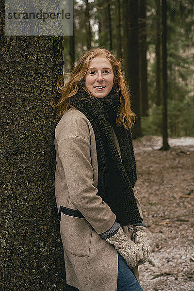Porträt einer selbstbewussten rothaarigen Frau mit Schal und Wollmantel  die sich an einen Baum im Wald lehnt