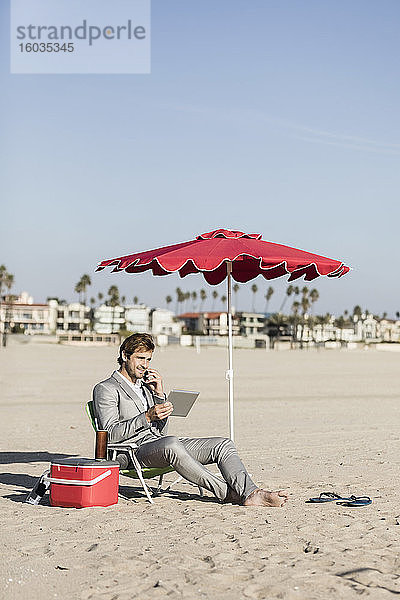 Barfüßiger Geschäftsmann mit Smartphone und digitalem Tablet arbeitet am Strand  Los Angeles  Kalifornien