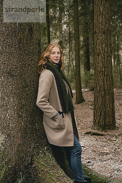 Porträt einer selbstbewussten rothaarigen Frau im Wollmantel  die sich im Wald an einen Baum lehnt