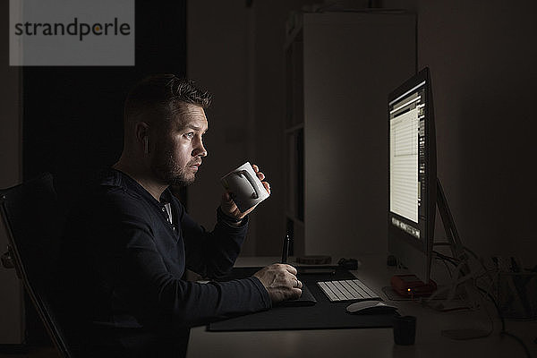 Mann mit Kaffee arbeitet spät am Computer in einem dunklen Raum