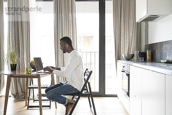 Lächelnder Mann sitzt am Tisch in einer modernen Wohnung und benutzt Laptop und Kopfhörer