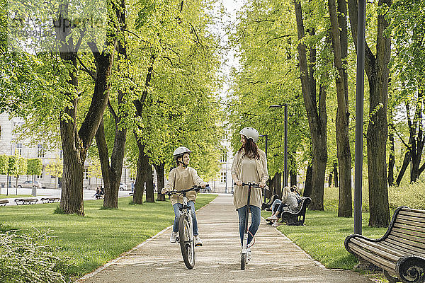 Mädchen auf dem Fahrrad  während die Mutter mit dem Elektroroller auf der Straße im Stadtpark fährt