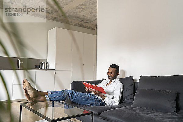 Lächelnder Mann sitzt auf der Couch in einer modernen Wohnung und liest ein Buch