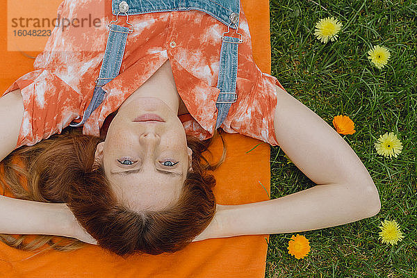 Entspannte junge Frau liegt mit den Händen hinter dem Kopf auf einer Picknickdecke bei Blumen im Hinterhof