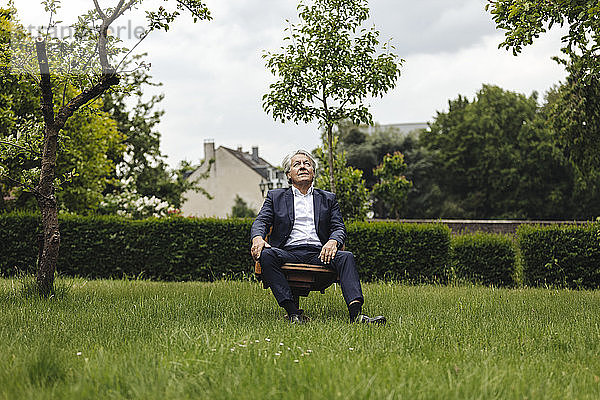 Älterer Geschäftsmann sitzt auf einem Stuhl in einem ländlichen Garten und schaut nach oben