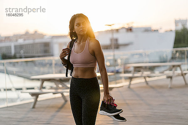 Nachdenkliche Sportlerin mit Schuhen in der Hand  während sie bei Sonnenuntergang am Pier steht