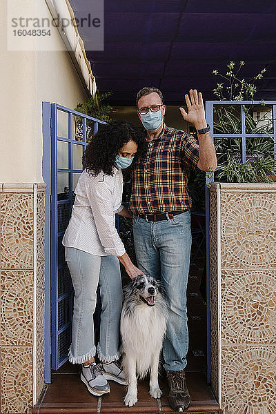 Ein Mann mit Maske winkt mit der Hand  während eine Frau einen Hund betrachtet
