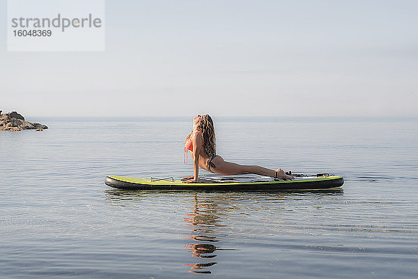 Frau übt nach oben gerichteten Hund Position auf Paddleboard über Meer gegen den Himmel