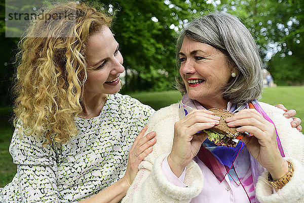 Lächelnde blonde Frau betrachtet ältere Mutter beim Essen eines Sandwiches im Park
