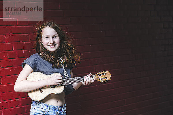 Lächelndes Mädchen spielt Ukulele  während sie gegen eine Backsteinmauer in der Stadt steht