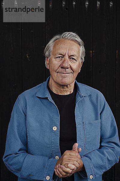 Porträt eines selbstbewussten älteren Mannes  der an einer Holzwand steht
