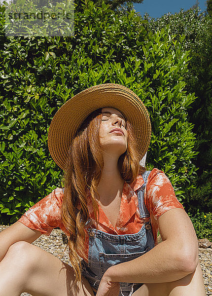 Schöne rothaarige Frau mit geschlossenen Augen sitzen gegen Pflanzen im Hinterhof auf sonnigen Tag