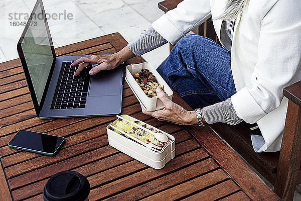 Mittelteil eines älteren weiblichen Berufstätigen  der eine Lunchbox hält und einen Laptop im Garten benutzt