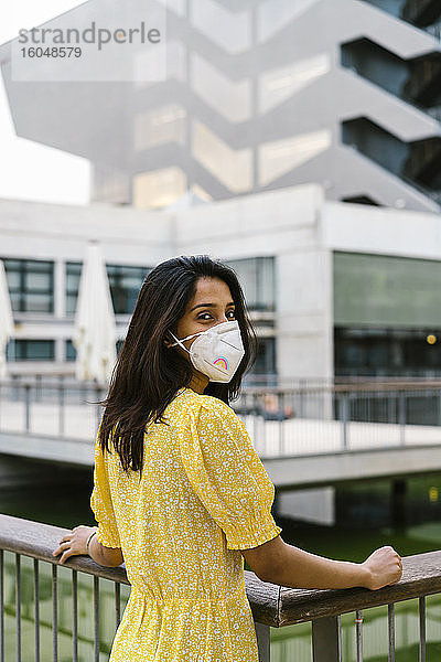 Junge Frau trägt Gesichtsmaske  während sie auf einem erhöhten Gehweg in der Stadt steht