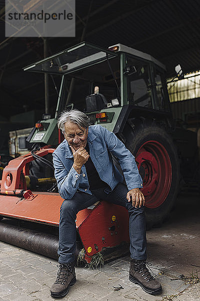 Lächelnder älterer Mann auf einem Bauernhof  der auf einem Traktor in einer Scheune sitzt