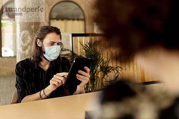 Junger Mann mit Maske  der in einem Restaurant sitzt und ein digitales Tablet benutzt