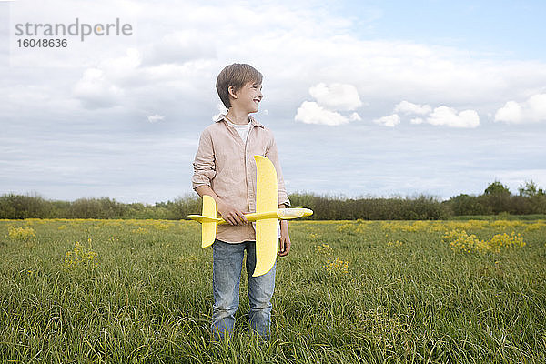 Lächelnder Junge  der ein Spielzeugflugzeug in der Hand hält und auf einem Rapsfeld in den Himmel schaut