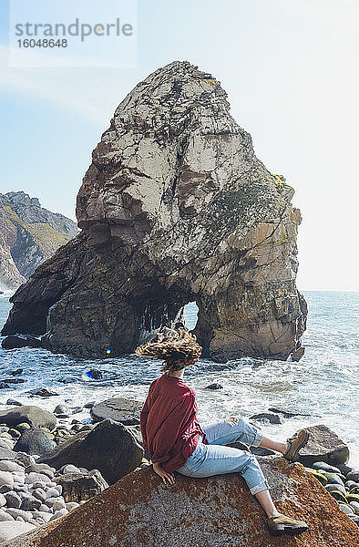 Sorglose Frau schüttelt den Kopf  während sie auf einem Felsen am Strand von Ursa sitzt  Portugal