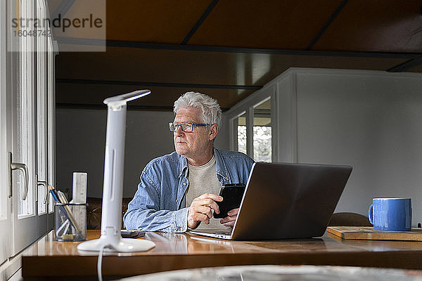 Nachdenklicher älterer Mann hält Smartphone mit Laptop auf dem Tisch zu Hause