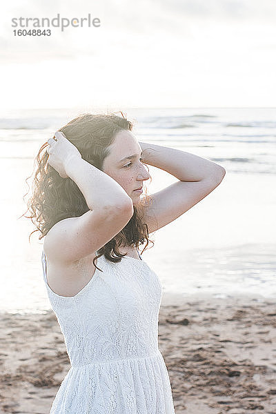 Nachdenkliche junge Frau in weißem Kleid mit Händen im Haar am Strand stehend