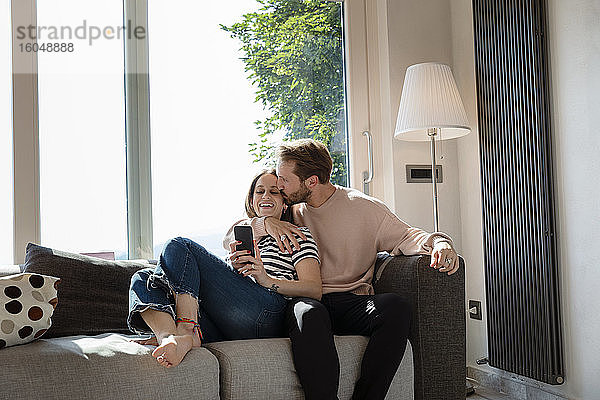 Mann küsst Frau beim Entspannen auf dem Sofa vor dem Fenster im Wohnzimmer