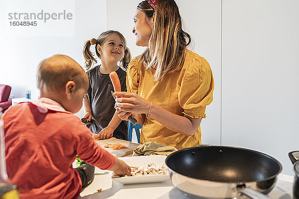 Lächelnde Mutter und Tochter schneiden Karotten  während das kleine Mädchen auf der Kücheninsel spielt