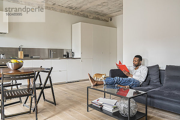 Mann sitzt auf Couch in moderner Wohnung und liest ein Buch