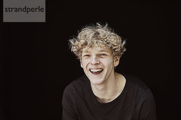 Porträt eines lachenden jungen Mannes mit lockigem blondem Haar vor schwarzem Hintergrund