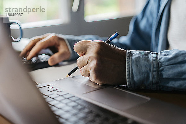 Ausgeschnittenes Bild eines älteren Mannes  der einen Taschenrechner und einen Laptop benutzt  während er zu Hause mit einem Bleistift schreibt